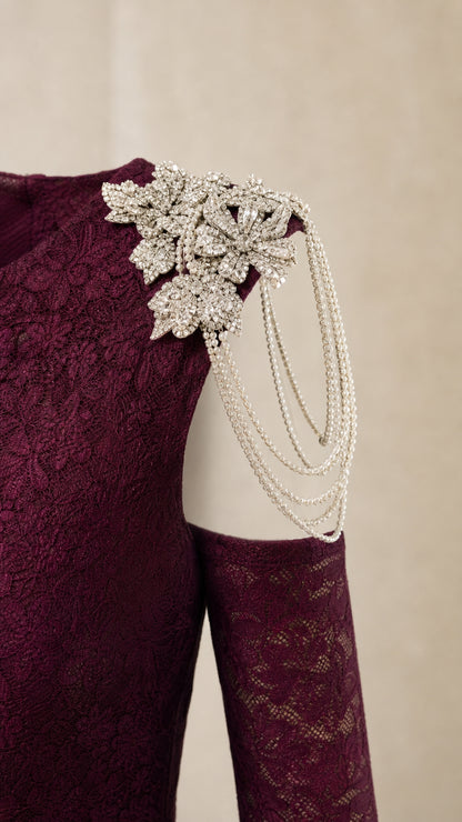 Close-up detail of crystal floral brooch with cascading chain trim on textured burgundy gown shoulder
